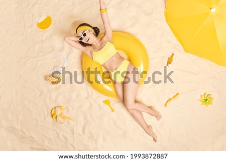Overhead shot of positive upbeat brunette young Asian woman wears sunglasses yellow swimsuit keeps arm raised has happy mood lies relaxed on swimring sunbathes at white sand after swimming in sea