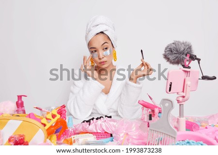 Serious Asian woman gies tips to followers about skin care and applying makeup holds cosmetic brush applies patches under eyes for rejuvenation records live stream video online uses beauty products