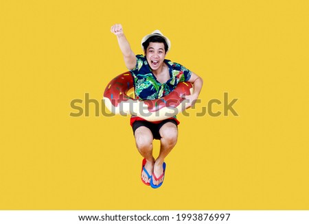 Young attractive Asian man wearing green and blue Hawaiian shirt and donut swim ring around his waist jumping against yellow background. Concept for holiday beach vacation.