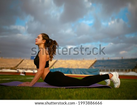 Fitness Girl Doing Leg Workout On Yoga Mat At Outdoor Stadium. Fit Woman With Strong Muscular Body In Fashion Sporty Outfit Doing Exercise Against stadium background. Active Lifestyle For Urban People