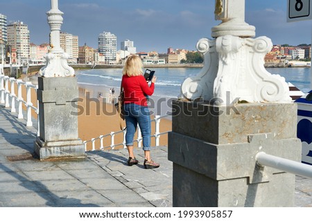 Woman in a red sweater and blue jeans photographs the beach of San Lorenzo in Gijon with her mobile phone