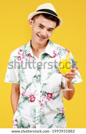 Young attractive Asian man in white Hawaiian shirt holding a glass of orange juice against yellow background. Concept for beach vacation holiday.