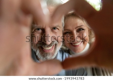 Crop close up portrait of happy mature man woman make heart love hand gesture sign pose for selfie together. Smiling old couple spouses have fun take self-portrait picture show family care bonding.