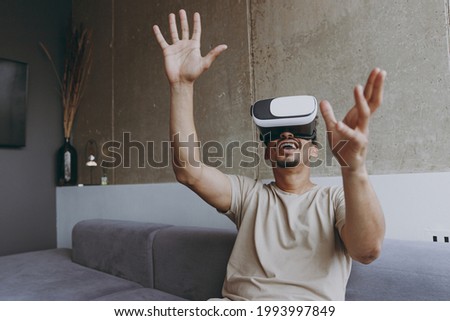 Happy impressed cool young african american man 20s wear beige t-shirt sit on grey sofa indoors apartment watch in vr headset pc gadget play game with outstretched hand rest on quarantine stay home