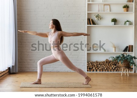 Peaceful focused girl standing in Warrior Two exercise, Virabhadrasana pose, doing yoga or pilates exercise wearing light pink sportswear. No stress concept. Healthy woman balance harmony.