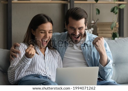 Happy emotional positive young family couple woman man looking at computer screen, celebrating online lottery giveaway betting auction win, feeling overjoyed together at home, internet success concept Royalty-Free Stock Photo #1994071262