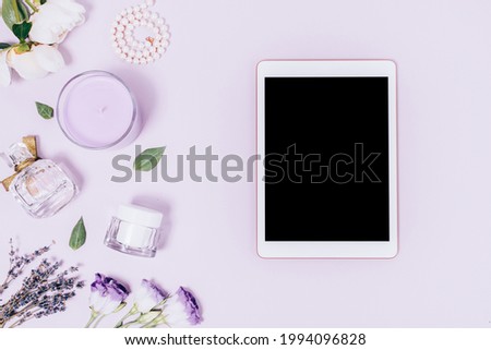 Feminine workplace, tablet pc with blank screen on lilac table next to flowers, perfume, scented candle and pearl necklace, flat lay.