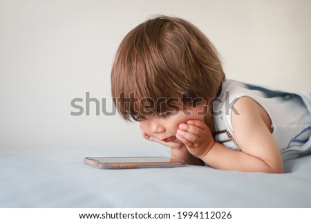 Cute baby lying on bed looking on smartphone. Mixed race Asian-German child watching cartoon or application on mobile phone at home. Kid learning with technology.