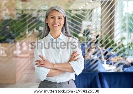 Happy smiling confident middle aged Asian older senior female leader businesswoman standing in modern office workplace looking at camera arms crossed. Business successful executive concept. Portrait. Royalty-Free Stock Photo #1994125508