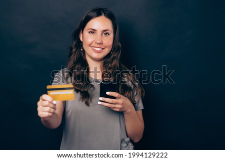 Photo of young smiling woman showing yellow credit card and using smartphone over black background.