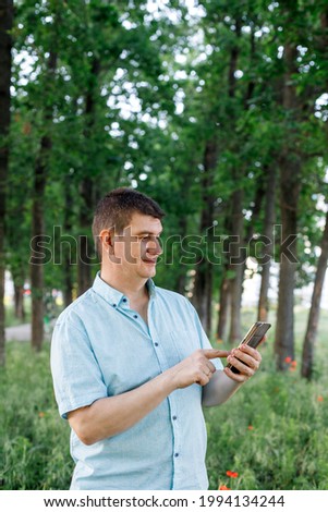 A brunette man in a blue short-sleeved shirt stands on the grass in an oak alley