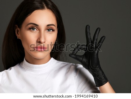 Portrait of attractive young woman doctor dentist in white uniform and latex gloves showing ok good sign with fingers over dark background. Dentist, stomatologist, orthopedist concept