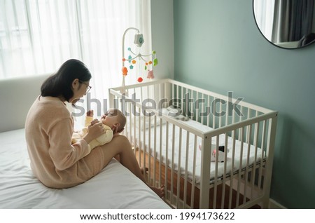 Beautiful young mother embracing and playing with her baby son in bedroom. Asian mom sitting on bed holding newborn child hands. Happy mother’s day.