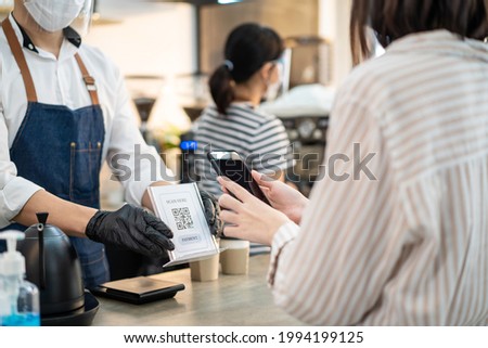 Close up of Asian woman customer make a quick and easy contactless payment in cafe during Covid19 Pandemic. Young girl use smartphone Scanning QR code to pay for service and drink from barista waiter. Royalty-Free Stock Photo #1994199125