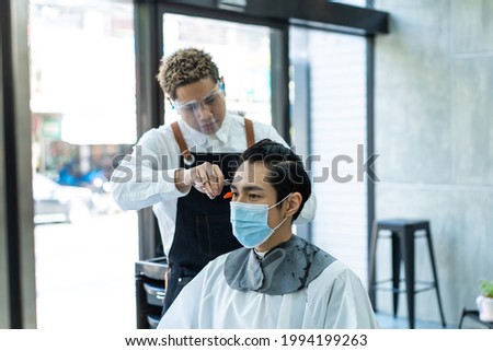 Asian professional male hairstylist combing and use scissors cutting young man customer's hair in salon. Hairdresser wearing protective face mask to prevent from coronavirus infection during pandemic. Royalty-Free Stock Photo #1994199263