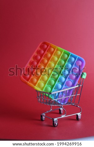 The multi-colored toy is popit simple dimple anti-stress in a basket of stroller shopping carts on a red background with a place for text copyspace. Shopping.