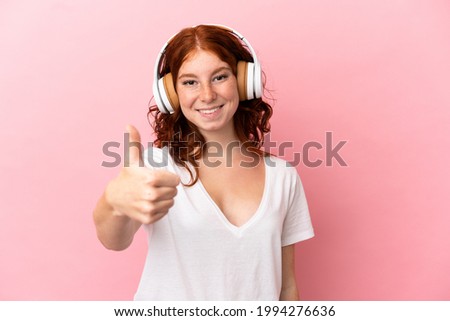 Teenager reddish woman isolated on pink background listening music and with thumb up