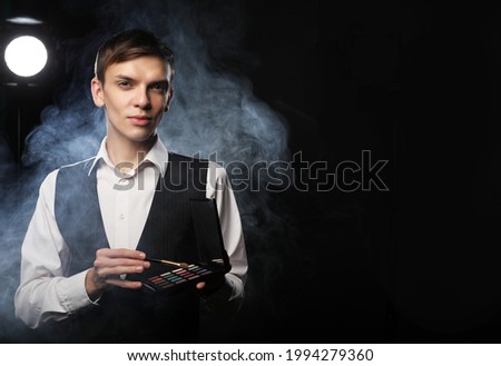 Portrait of attractive guy make-up artist holding in hand eye shadow palette over black background
