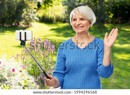 gardening, technology and old people concept - smiling senior woman taking picture by smartphone on selfie stick waving hand over summer garden background
