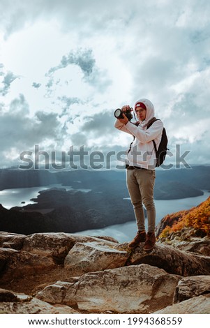 A young male photographer wearing a hoodie with a backpack and taking photos of hills with a DSLR camera