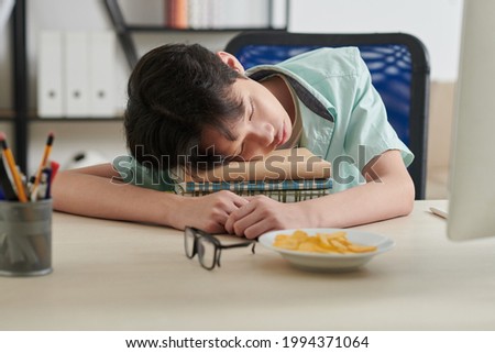 Tired teenage schoolboy sleeping on stack of books on his desk at home after studying online al day long
