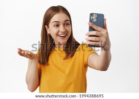 Image of excited surprised girl talking video call conference on smartphone, showing something to mobile camera, raising hand and smiling happy, white background