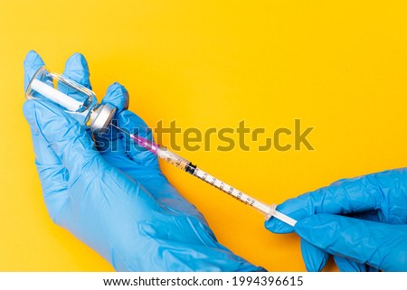 Hands in blue gloves holding syringe and stabbing to a bottle of a vaccine on orange background with copy space.