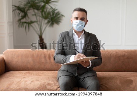 Professional Therapist Concept. Portrait Of Smiling Man Wearing Suit And Protective Medical Face Mask Sitting On Couch Sofa, Holding Clipboard With Pen And Writing, Posing And Looking At Camera