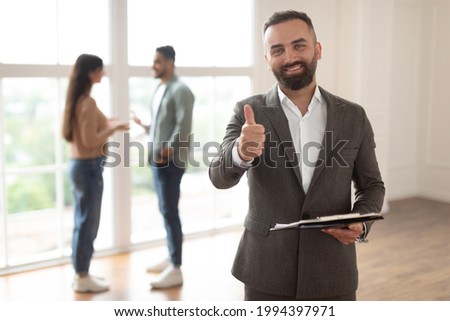 Successful Deal, House Ownership Concept. Portrait Of Smiling Professional Real Estate Agent In Suit Showing Thumb Up Sign Like Gesture, Selective Focus. Buyers Couple Talking In Blurred Background