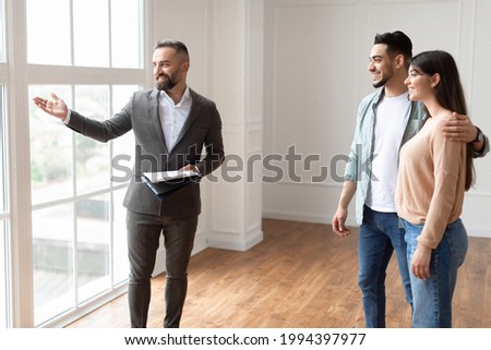 Housing Concept. Property Agent In Suit Showing View From Big Floor-to-Ceiling French Windows To Future Homeowners, Making Tour In New Empty Apartment To Young Family, Husband Embracing Wife