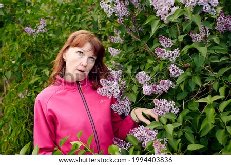 Photo of a caucasian woman in a pink jacket outdoors on the background of a lilac bush posing