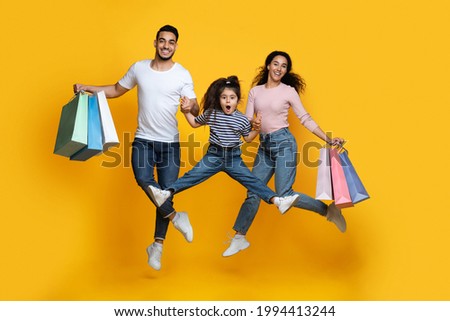 Seasonal Sales Concept. Happy Middle Eastern Family Of Three Jumping With Shopping Bags Over Yellow Studio Background, Arab Parents And Little Daughter Having Fun Together, Full Length, Free Space