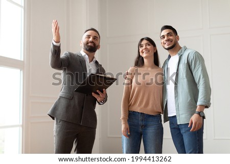 Portrait Of Male Realtor Or Architect Wearing Suit Showing New Empty House To Smiling Millennial Buyers, Pointing. Husband Embracing His Wife, Visiting Residential Building, Choosing Home For Family Royalty-Free Stock Photo #1994413262