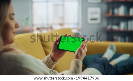 Beautiful Authentic Female Using a Smartphone with Green Screen Mock Up Display at Home Living Room while Lying on a Couch Sofa. She's Browsing the Internet and Checking Videos on Social Networks.