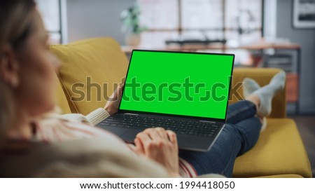 Beautiful Caucasian Specialist Working on Laptop with Green Screen Mock Up Display at Home Living Room while Lying on a Couch Sofa. Freelance Female Chatting Over the Internet on Social Networks.