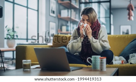 Ill Young Female Blowing Her Nose and Calling into Office with Video Phone Call from Cozy Living Room. Sick and Nauseous Woman in Warm Scarf is Using Laptop, Watching Movies. Royalty-Free Stock Photo #1994419313