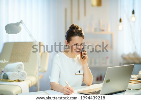 happy woman worker with laptop speaking on a smartphone in modern beauty salon. Royalty-Free Stock Photo #1994439218