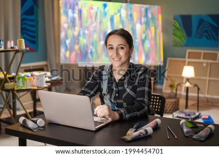 A smiling brunette sits at a desk in front of a laptop in art studio and displays paintings for sale, around her lies a brush, watercolor paints in a tube, in the background a multi-colored painting