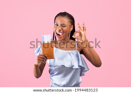 Positive black woman holding passport and plane tickets, showing okay gesture and winking over pink studio background. Millennial lady going on summer vacation, traveling around world