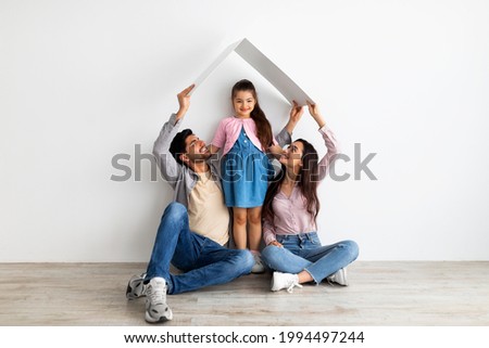 Housing for young family concept. Young eastern father, mother and daughter under symbolic roof dreaming of new home, sitting on floor over light wall Royalty-Free Stock Photo #1994497244