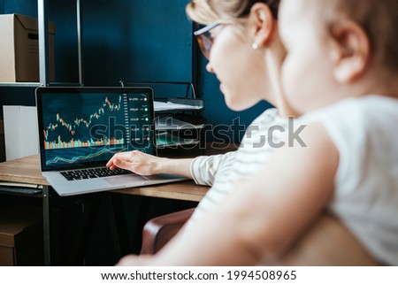 Handsome woman with her cute baby investing in world stock market, using her laptop and online trading soft, working from home