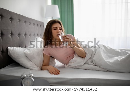 Young Caucasian woman drinking clear mineral water from glass while lying in bed at home. Lovely millennial lady staying hydrated, taking care of herself after sleep. Healthy living concept Royalty-Free Stock Photo #1994511452