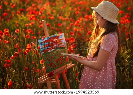 Little girl is standing in the field of red poppies and painting on the canvas placed on a drawing stand. Be a part of learning outside of the school in the nature park. Creative hobby