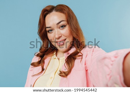 Close up young smiling fun redhead chubby overweight woman 30s in pink shirt doing selfie shot on mobile phone look camera isolated on pastel blue background studio portrait. People lifestyle concept