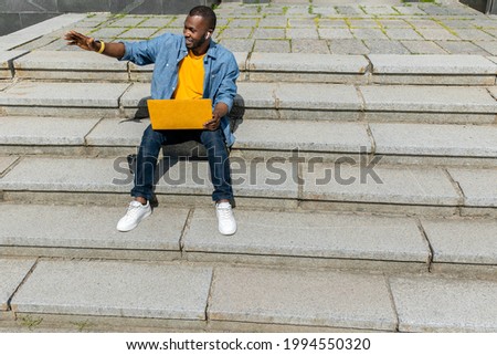 Joyful afro american freelancer businessman using yellow laptop outdoors,sitting on stairs in the park, talking online with colleagues or family. Handsome male taking break after online remote meeting