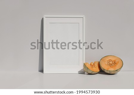 Modern tropical summer still life scene. Empty vertical white wooden picture frame mockup.  Cut orange cantaloupe melon fruit, long shadows. Beige table and wall background. Art display concept.