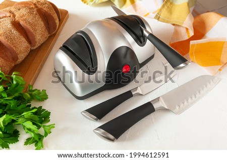 Electric knife sharpener. The plastic body is gray-black. A kitchen knife in the sharpener. In the foreground are kitchen knives. On the back there is bread and greens. Light background. Royalty-Free Stock Photo #1994612591