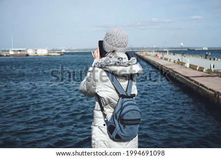 Girl with a backpack and hat and jacket on a background of the sea. Woman takes pictures by phone of ships at sea. Winters beautiful sea.