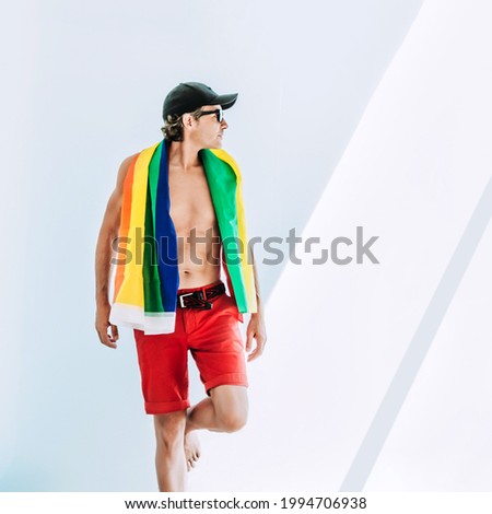 an athletic middle-aged man with a pride flag hanging from his neck looks sideways with a cap and has a bare torso. lgtb concept, equality and respect.