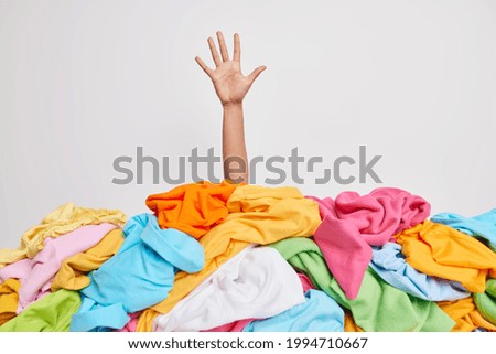 Unrecognizable human raises arm reaches out heap of colorful unfolded clothes busy doing wardrobe cleaning isolated over white background. Woman buried under cluttered clothing items. Decluttering Royalty-Free Stock Photo #1994710667